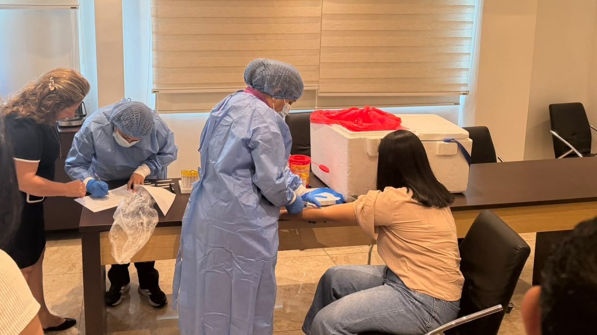 Dos profesionales de Simelab con bata, gorro y guantes realizan la toma de muestra sobre una mesa montada en la sala de reuniones de una empresa, junto a gradillas con tubos y un cooler de transporte.