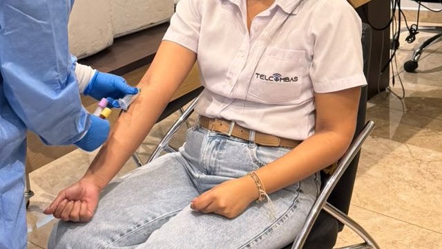 Primer plano de la toma de muestra: la mano enguantada de una profesional de Simelab sostiene el tubo mientras se llena, apoyada en el brazo de una trabajadora sentada.